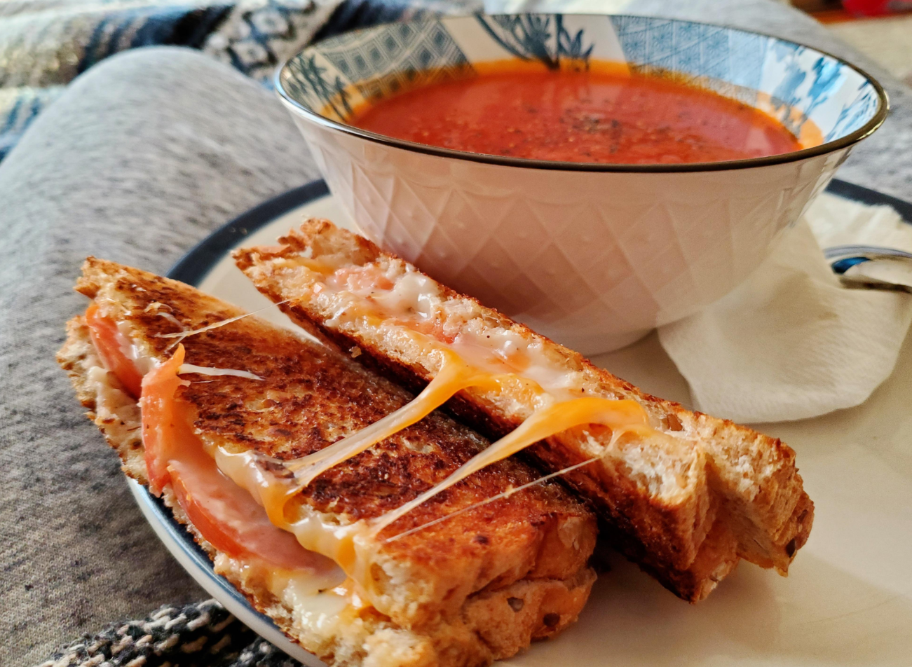 A bowl of tomato soup sits next to a plate with a grilled cheese sandwich cut in half, melted cheese stretching between the slices—one of those classic dinners served on a cozy surface with a patterned blanket in the background.