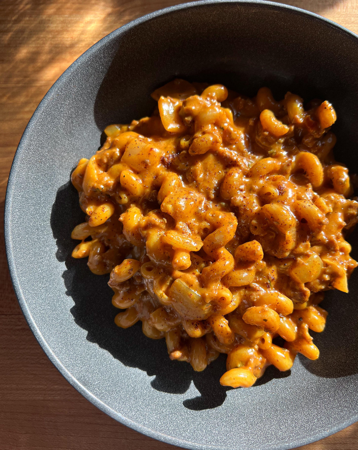 A dark bowl filled with creamy macaroni pasta in a rich, orange-yellow sauce, mixed with ground meat and onions, shown under natural sunlight—a comforting nod to classic dinners.