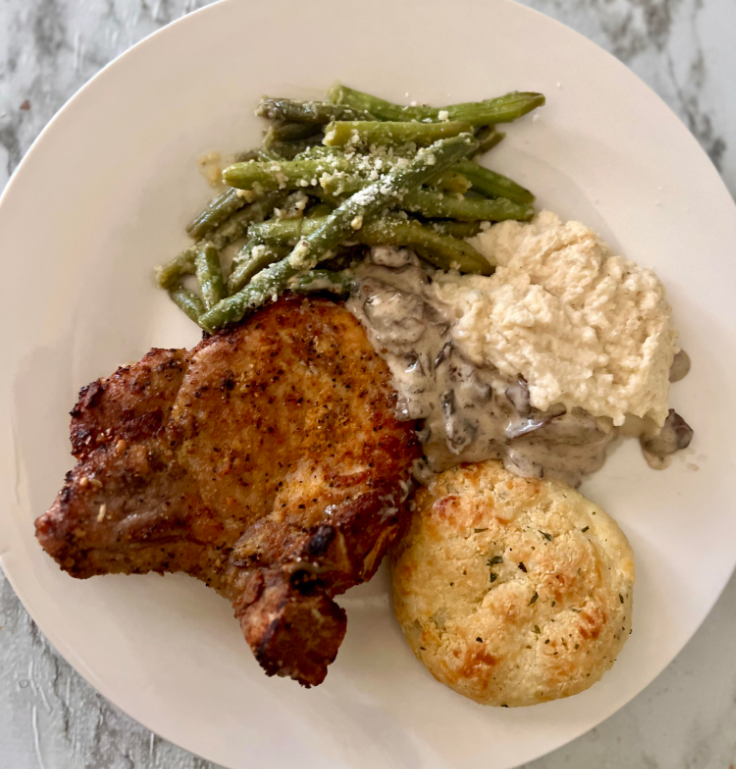 A white plate with a seasoned pork chop, green beans topped with grated cheese, creamy mashed potatoes with gravy, and a golden biscuit—this meal is the perfect example of classic dinners done right.