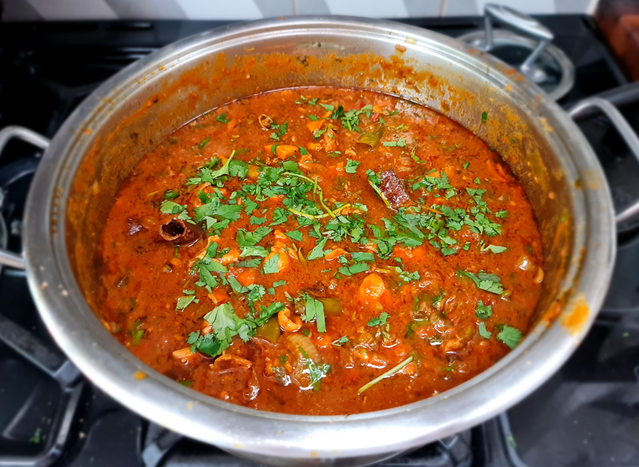 A large metal pot filled with rich, red curry—a staple of classic dinners—garnished with chopped fresh cilantro sits on a stove, with visible pieces of meat and spices in the sauce.