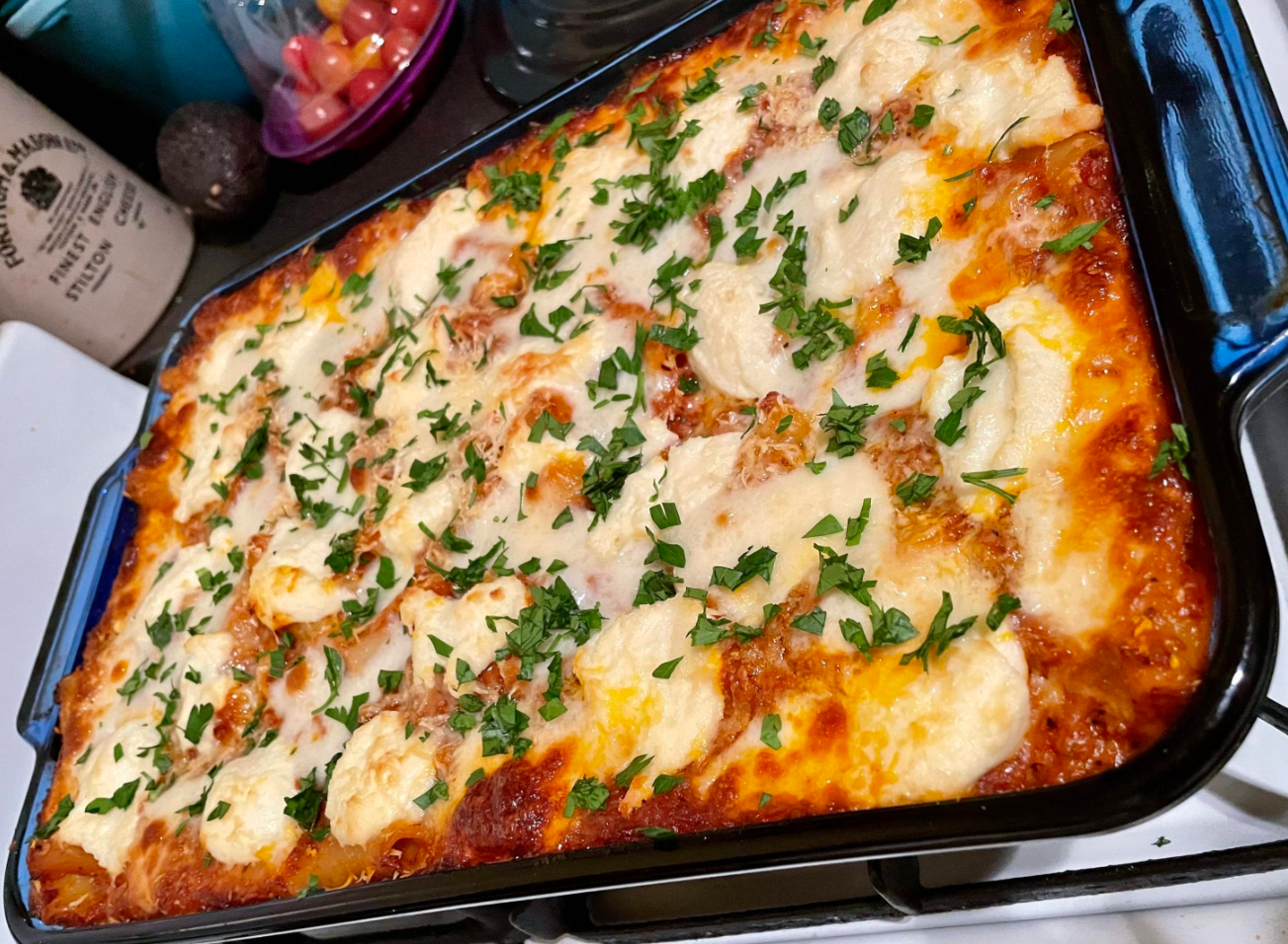 A freshly baked lasagna in a rectangular pan, topped with melted cheese, tomato sauce, and sprinkled with chopped parsley. This golden and bubbly dish is a favorite among classic dinners, sitting on a stovetop with kitchen items in the background.