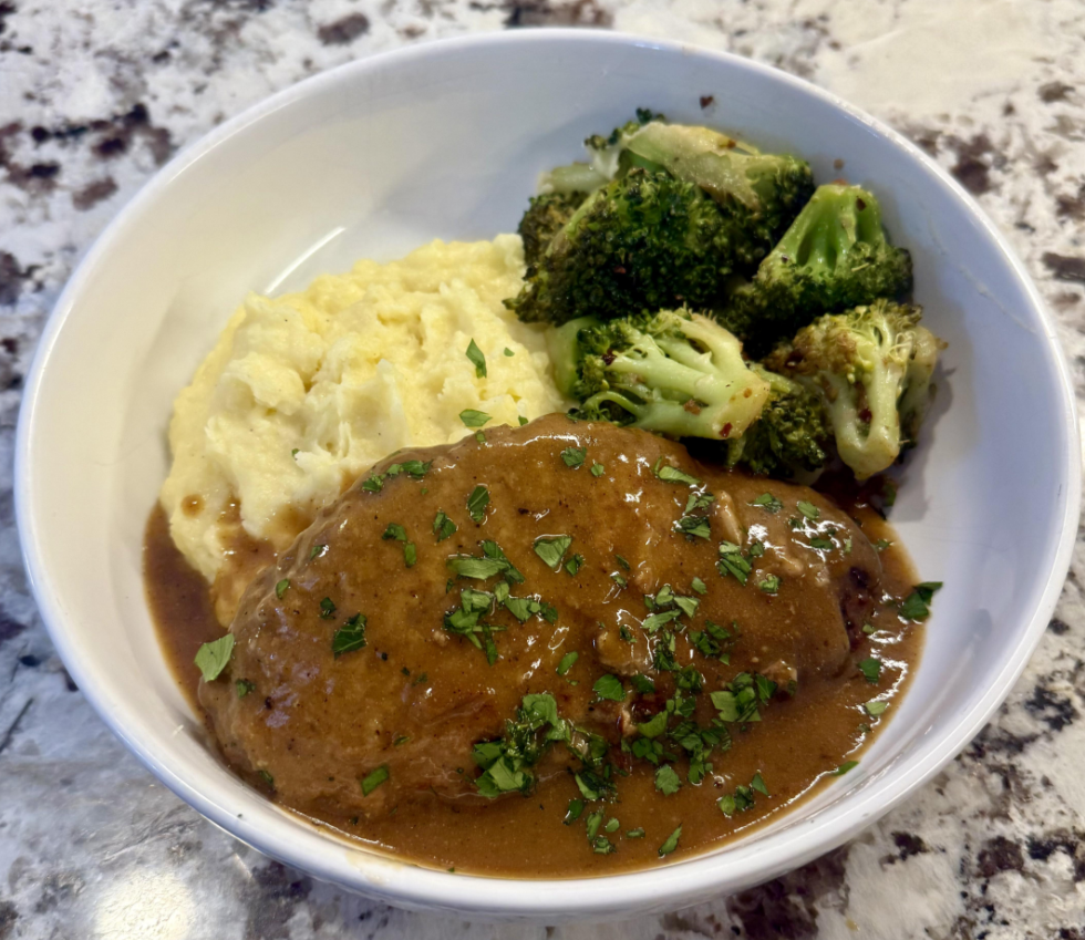 A bowl with mashed potatoes, steamed broccoli, and a piece of meat topped with brown gravy and chopped herbs, placed on a marbled countertop—a perfect example of classic dinners.