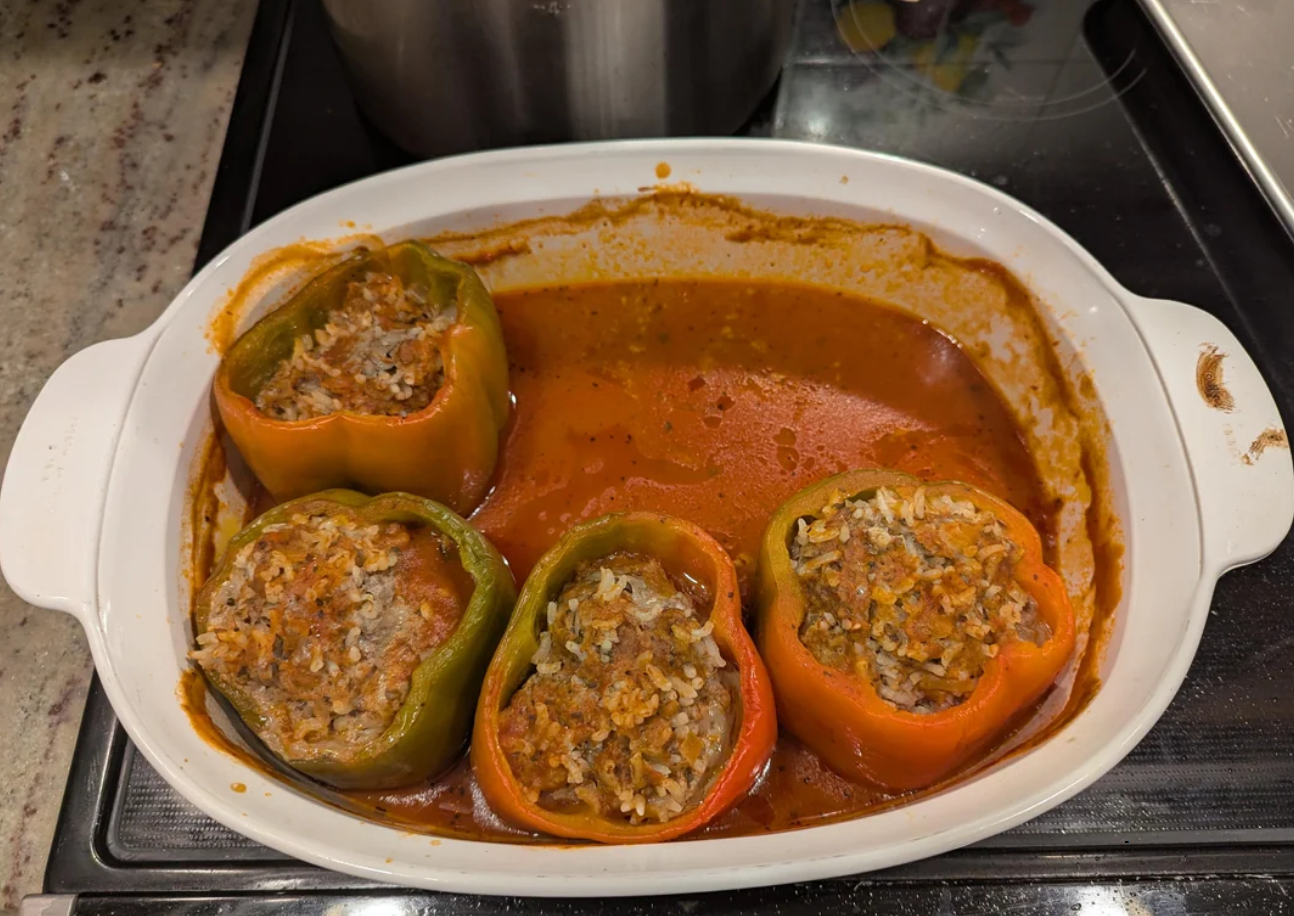 Four stuffed bell peppers, two green and two orange, are filled with a rice and meat mixture and baked in tomato sauce—a comforting option for classic dinners. The dish is served in a white pan set on the stovetop.
