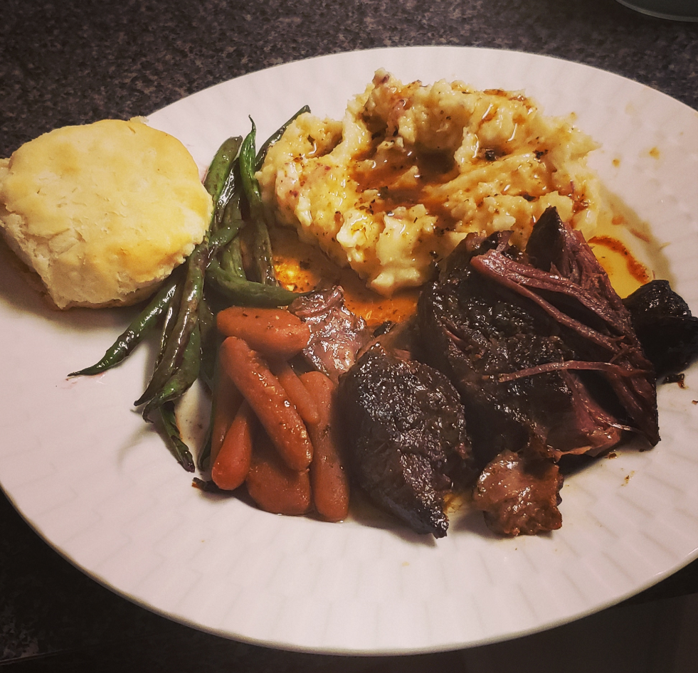 A white plate with mashed potatoes and gravy, pot roast with sauce, glazed baby carrots, green beans, and a biscuit on a speckled countertop—one of those classic dinners everyone loves.