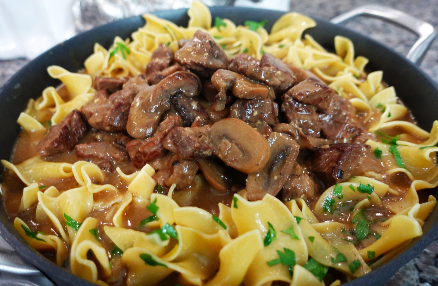 A skillet filled with beef stroganoff, one of the classic dinners, featuring pieces of beef, sliced mushrooms, and creamy sauce served over wide egg noodles, garnished with chopped parsley.