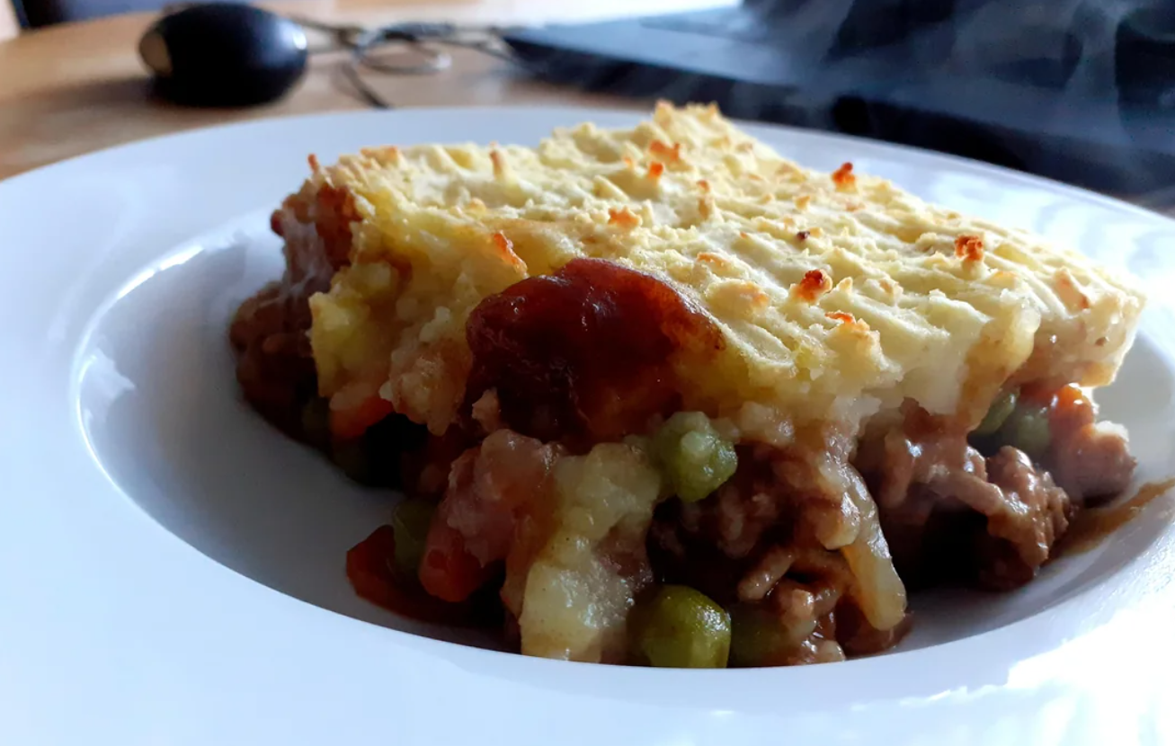 A close-up of a serving of shepherd’s pie, one of the classic dinners, on a white plate, showing mashed potatoes atop a savory meat, pea, and vegetable filling. A computer mouse and keyboard are blurred in the background.