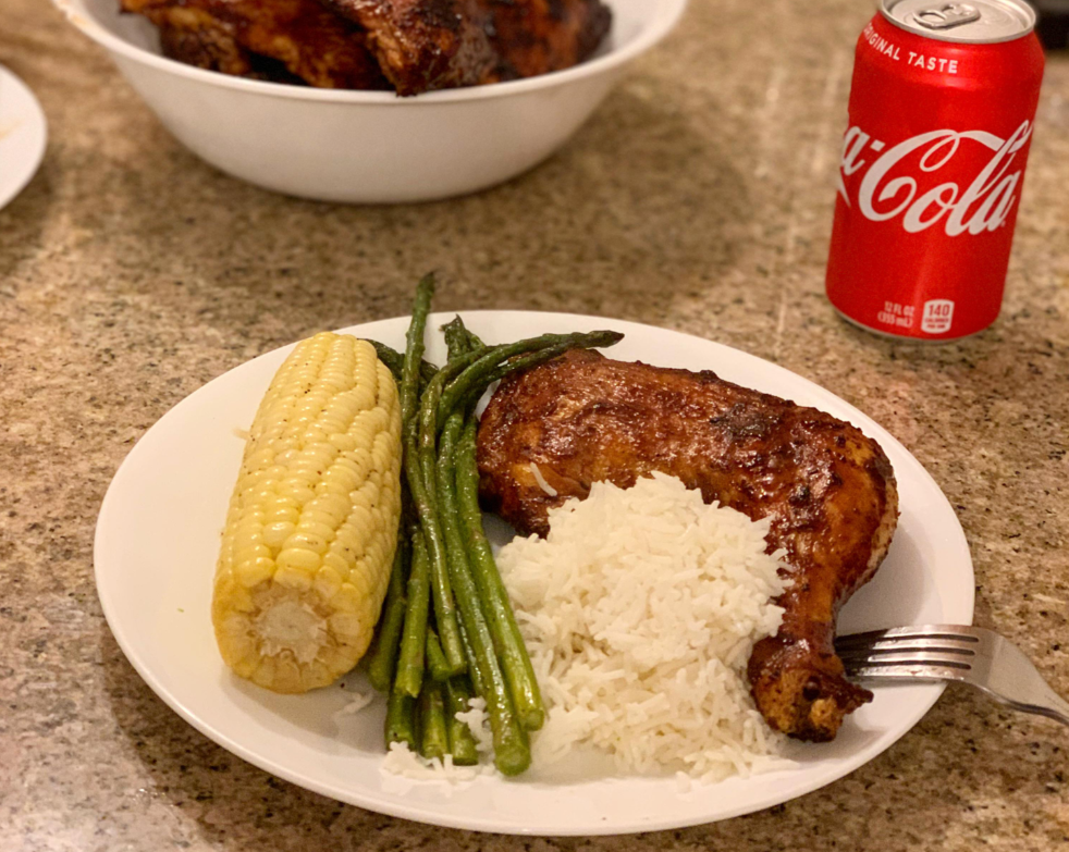 A plate of food with a grilled chicken leg, white rice, green beans, and corn on the cob sits on a counter next to a can of Coca-Cola—a perfect example of classic dinners. A white bowl with more chicken is in the background.