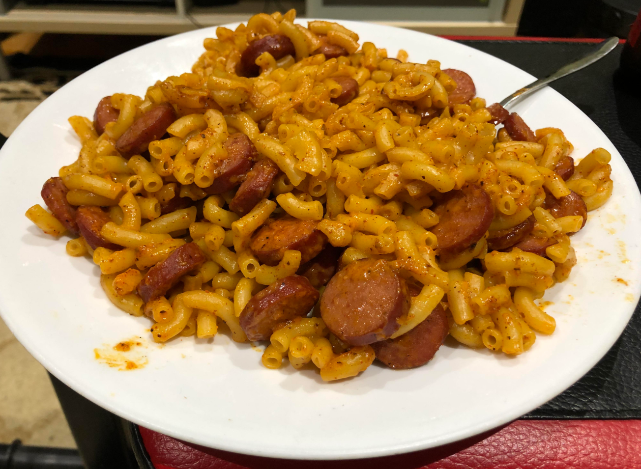 A white plate piled with macaroni and sliced sausage mixed in a reddish-orange sauce, with a fork resting on the plate. This hearty and flavorful meal is a comforting choice for fans of classic dinners.