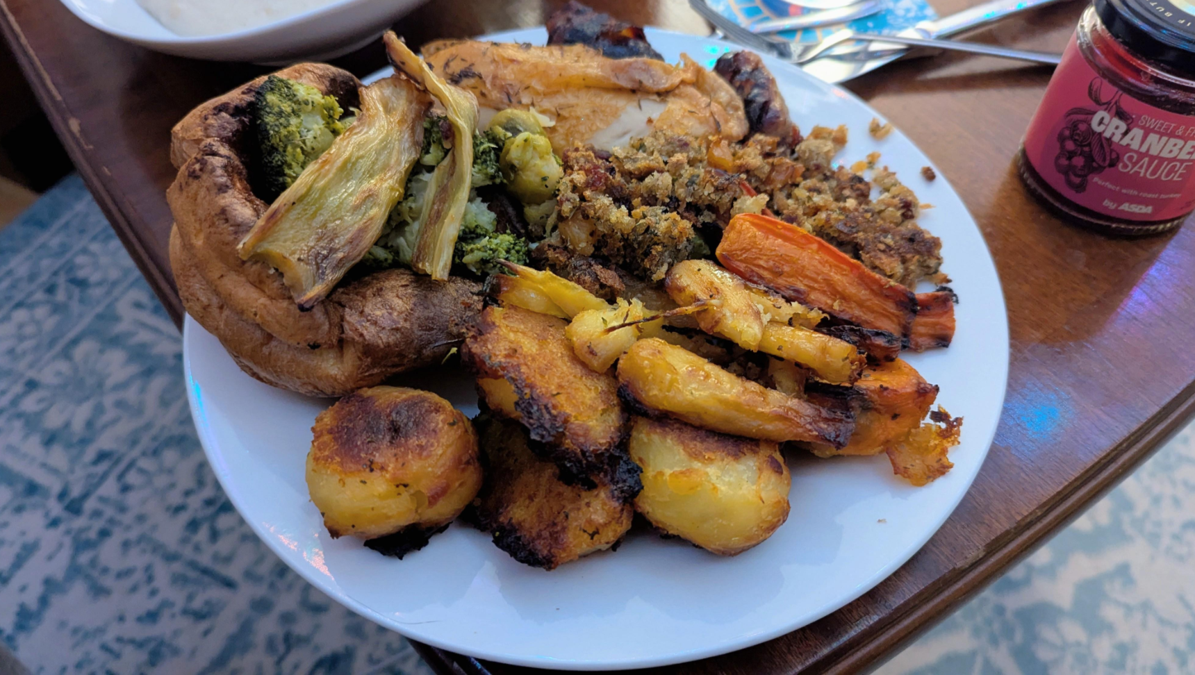 A plate of roast dinner with Yorkshire pudding, roasted potatoes, broccoli, carrots, stuffing, and parsnips sits on a table next to a jar of cranberry sauce—a classic favorite among middle class moms.