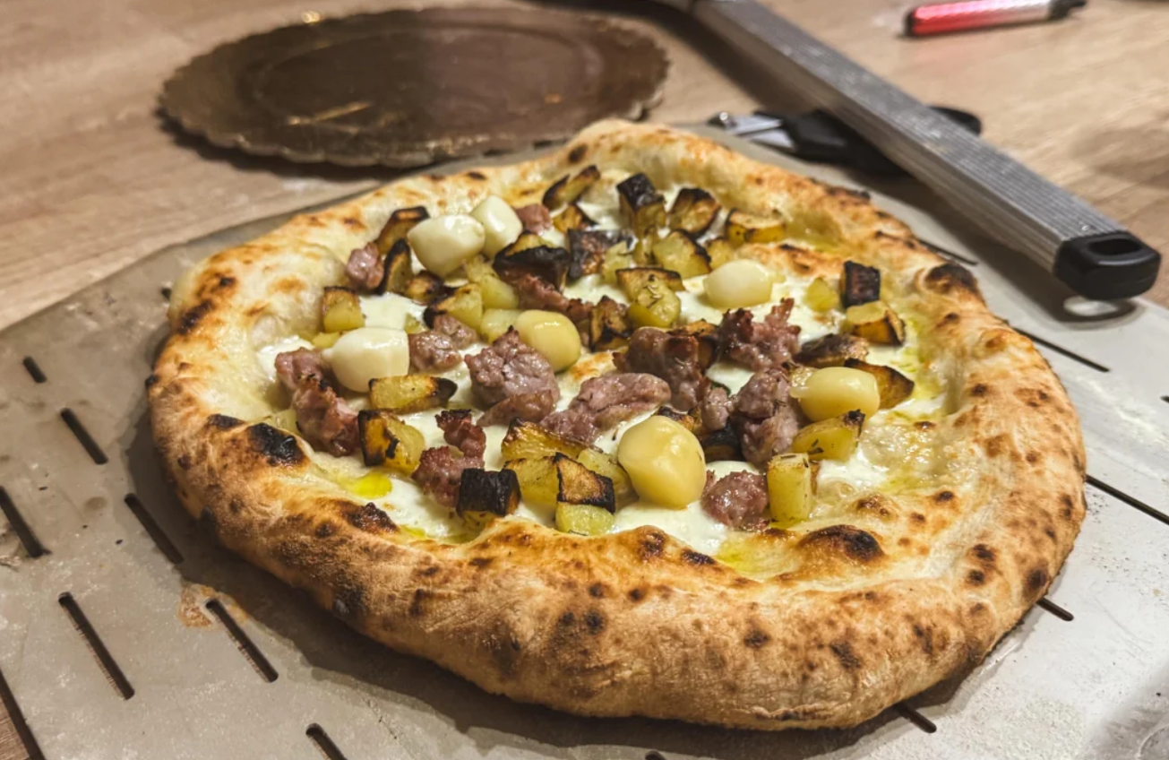 A freshly baked pizza topped with browned potato cubes, sausage pieces, and melted cheese sits on a perforated pizza peel—an easy favorite among middle class moms—with kitchen tools visible in the background.