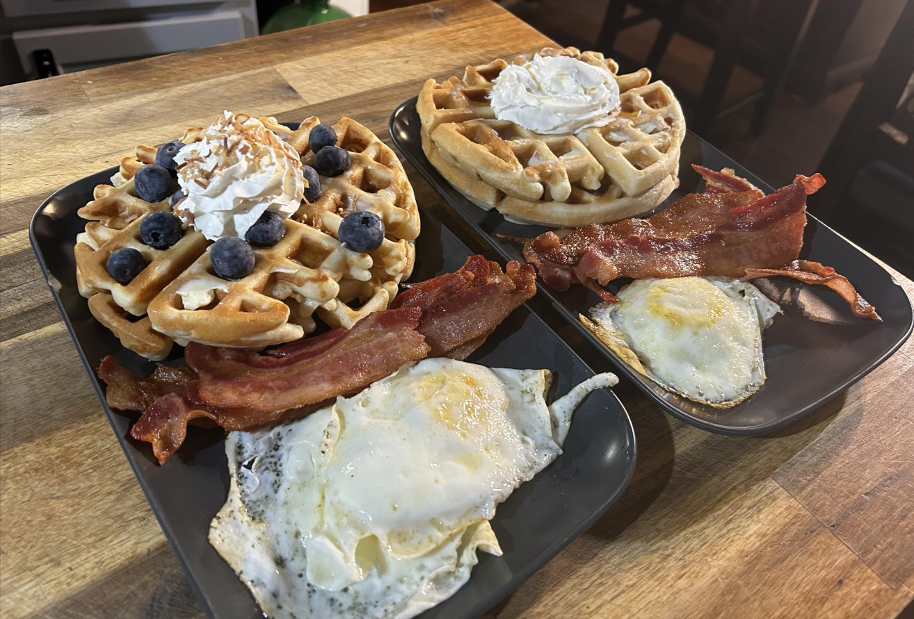 Two plates with breakfast food: each has two fried eggs, crispy bacon strips, and a large waffle topped with whipped cream. Perfect for middle class moms, one waffle is also garnished with blueberries and coconut flakes on a wooden table.