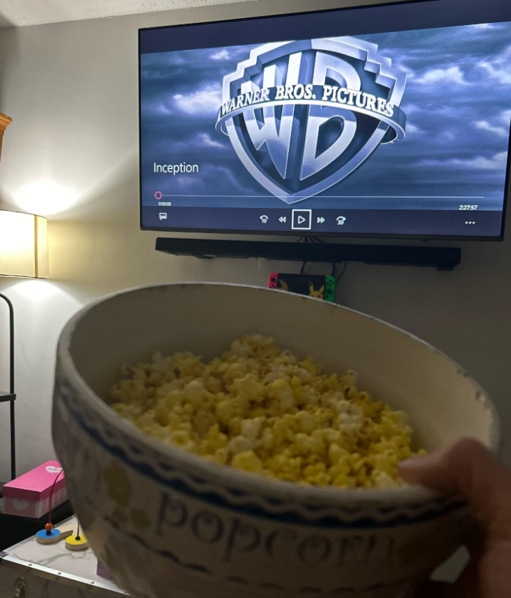 A hand holds a bowl of popcorn facing a TV displaying the Warner Bros. Pictures logo and "Inception," capturing a cozy movie night at home—a scene many middle class moms will recognize and enjoy.