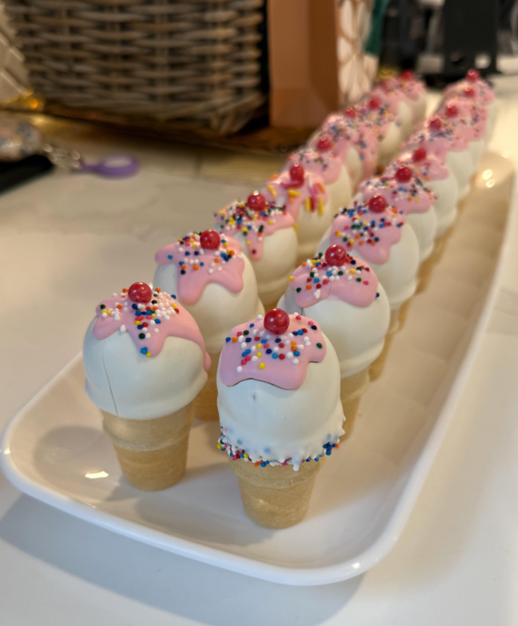 Mini ice cream cone treats with white coating, pink icing, rainbow sprinkles, and a red candy on top—perfect for middle class moms to serve at gatherings—arranged in neat rows on a white rectangular plate.