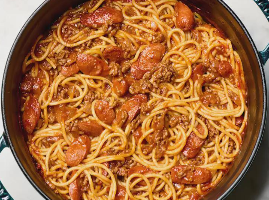 Cooked spaghetti with ground beef and slices of sausage in a rich red tomato sauce, just like middle class moms make, served in a dark round pot on a white surface.