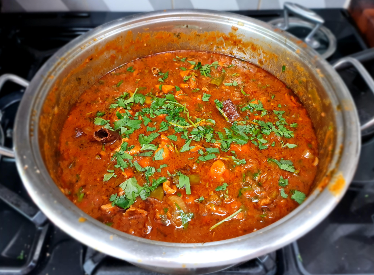 A stainless steel pot filled with rich, red curry garnished with fresh chopped cilantro sits on a stovetop—a comforting meal often enjoyed by middle class moms. Visible ingredients include chunks of meat and whole spices in the thick, aromatic sauce.