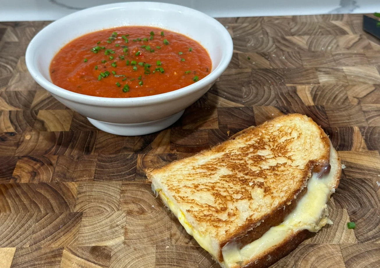 A comforting meal awaits: a bowl of tomato soup with chopped chives alongside a golden-brown grilled cheese, perfect for middle class moms seeking an easy yet hearty lunch, all served on a rustic wooden cutting board.