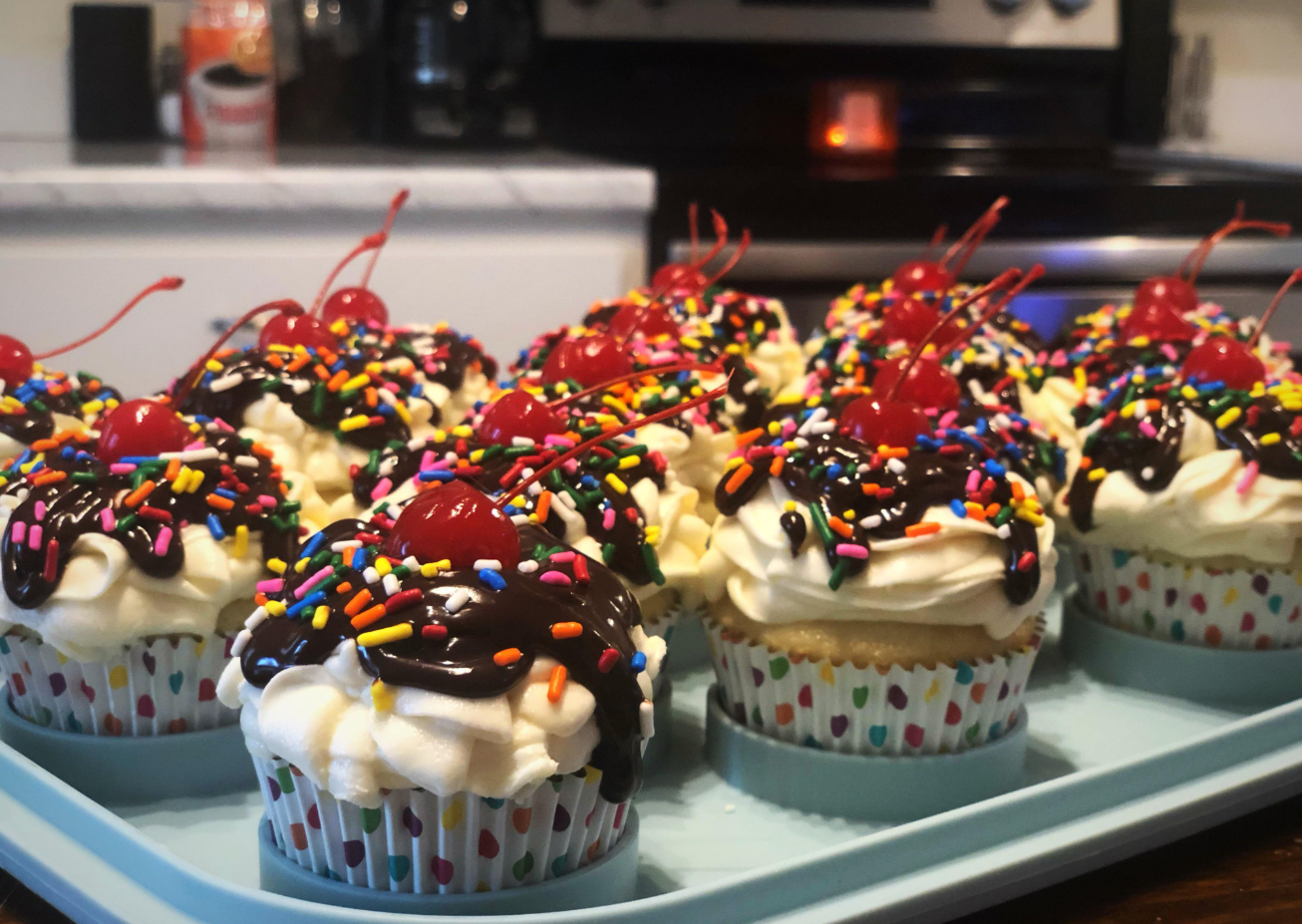 A tray of cupcakes with white frosting, chocolate drizzle, rainbow sprinkles, and a cherry on top sits on a kitchen counter—a sweet treat often enjoyed by middle class moms in their cozy homes.