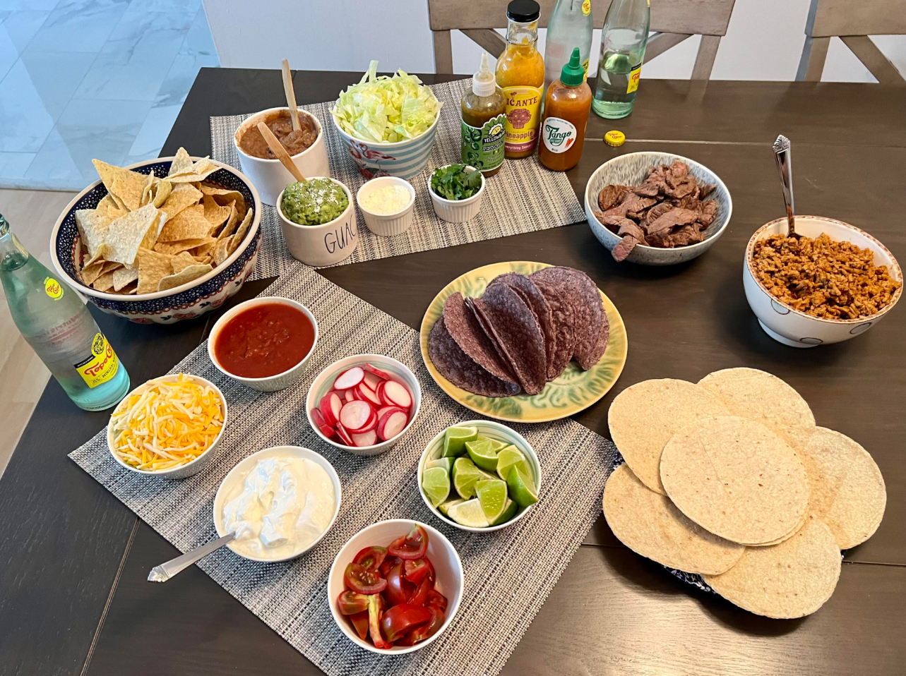A table set for taco night—perfect for middle class moms—with bowls of tortilla chips, shredded cheese, salsa, sour cream, guacamole, limes, radishes, grilled beef, rice, blue corn taco shells, corn tortillas, and hot sauces.