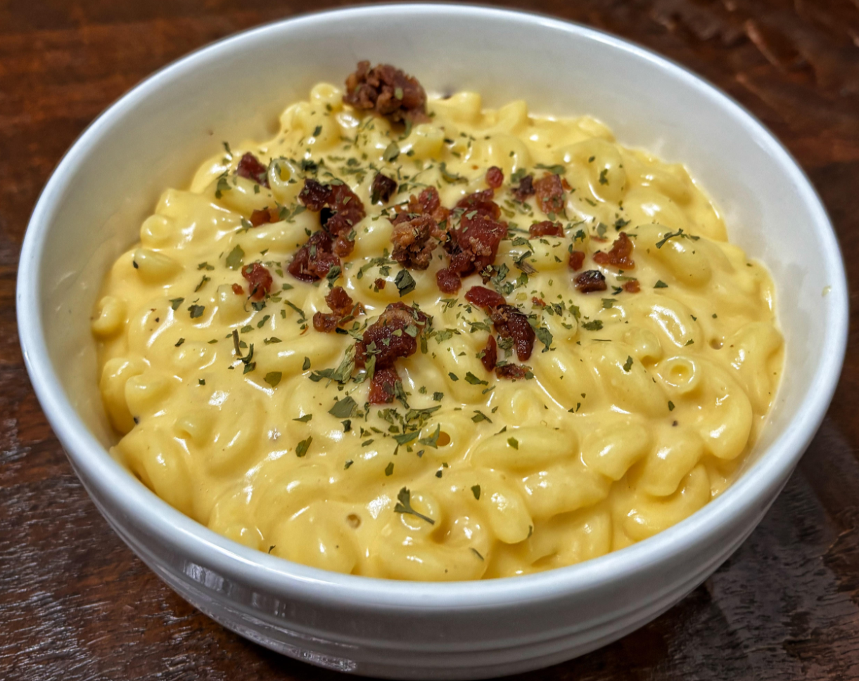A favorite among middle class moms, this white bowl filled with creamy macaroni and cheese, topped with crispy bacon pieces and sprinkled with herbs, sits on a dark wooden surface.