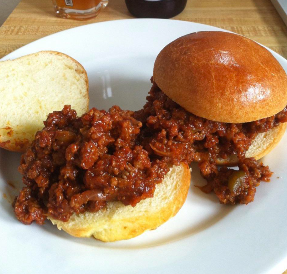 A white plate with two open-faced sloppy joe sandwiches, just like middle class moms might serve, showing seasoned ground beef filling on soft hamburger buns, set on a wooden surface.