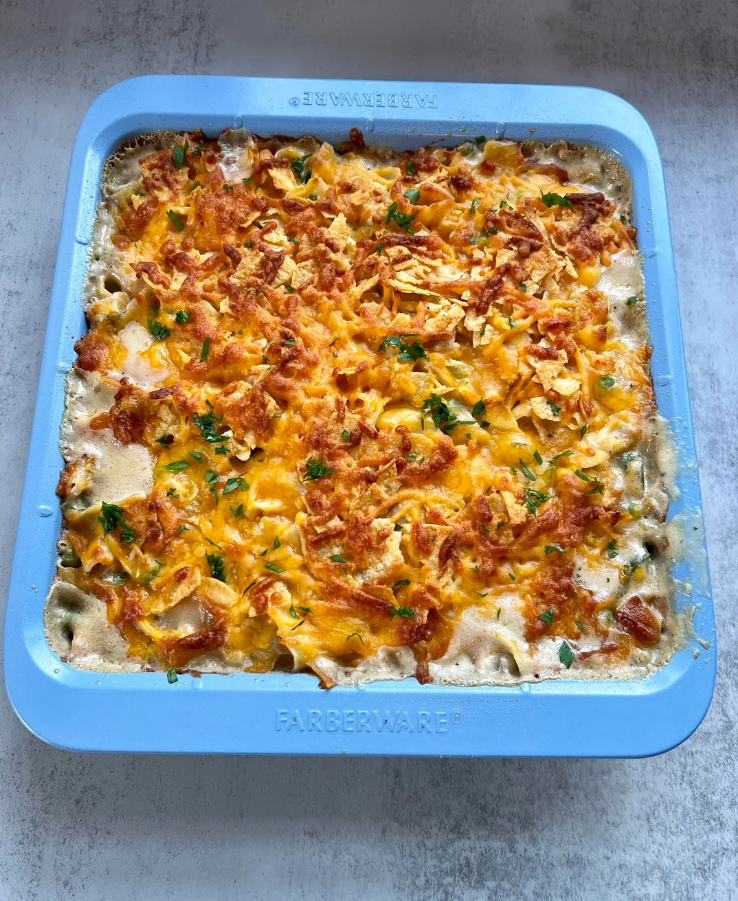 A square blue baking dish filled with a golden-brown casserole topped with melted cheese, crispy onions, and garnished with chopped herbs—a classic comfort dish loved by middle class moms—rests on a light-colored surface.