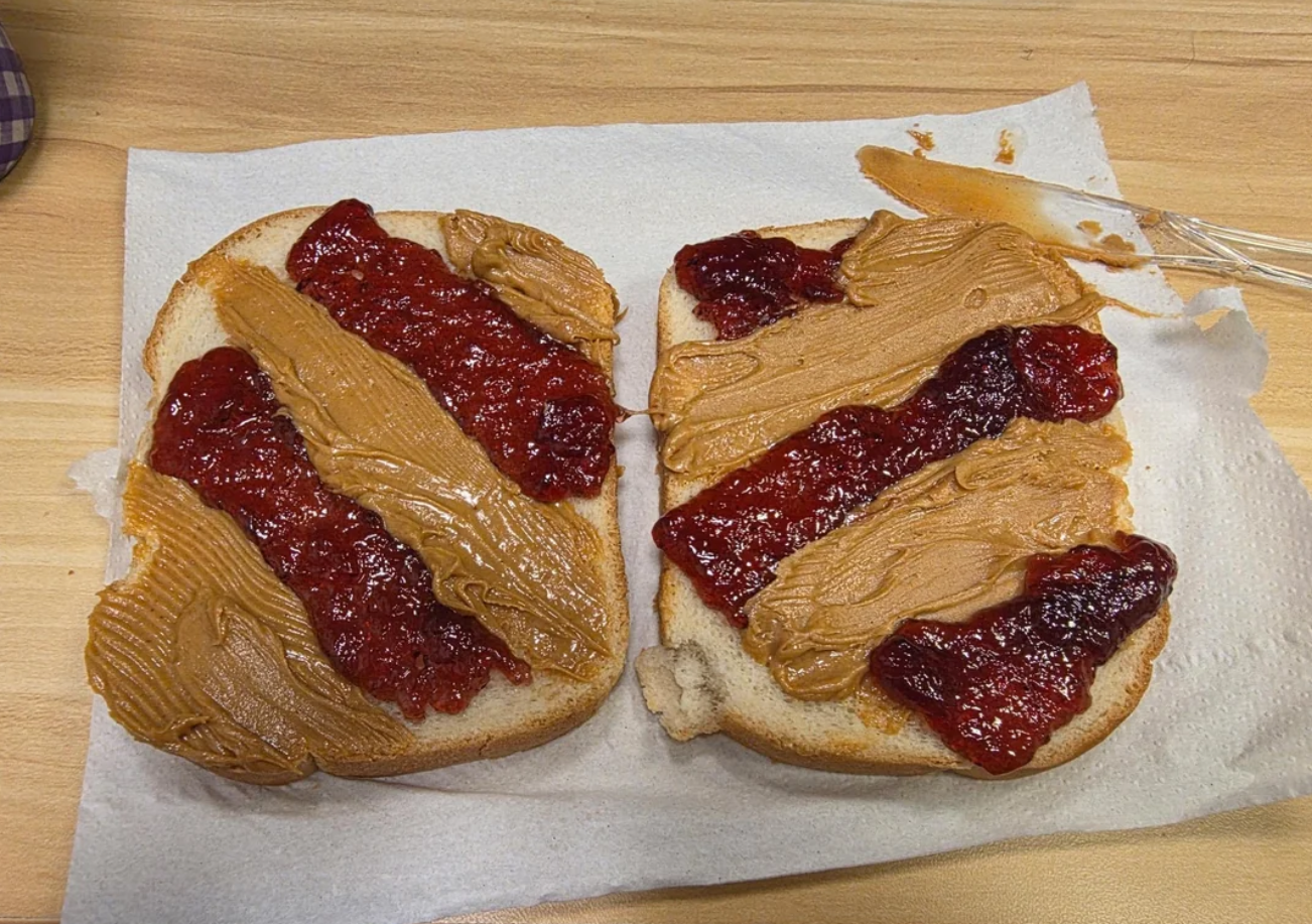 Two slices of white bread on a napkin, each spread with alternating diagonal stripes of peanut butter and grape jelly—just the kind of classic snack middle class moms love to make—sit beside a knife with some peanut butter and jelly residue.