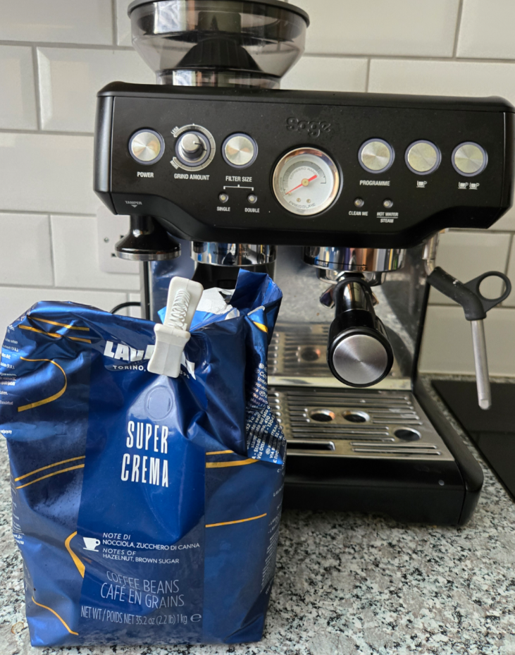 A black espresso machine sits on a kitchen counter with a blue bag of Lavazza Super Crema whole coffee beans placed in front of it. The background features white subway tiles.