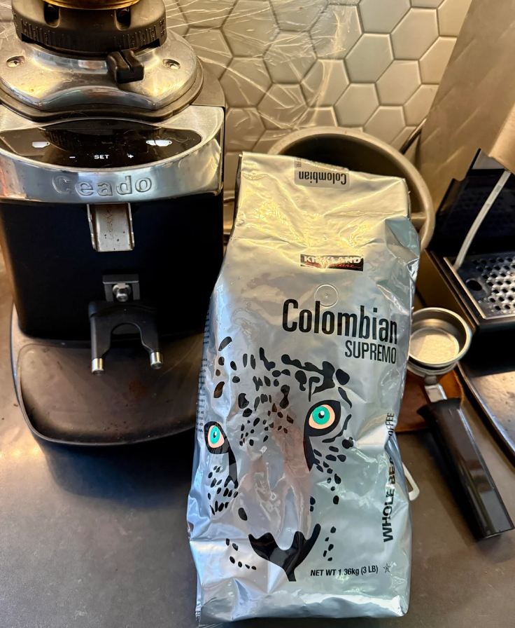 A silver bag of Kirkland Colombian Supremo coffee with a jaguar face design is on a kitchen counter next to a Ceado coffee grinder and a portafilter, with a honeycomb tile backsplash in the background.