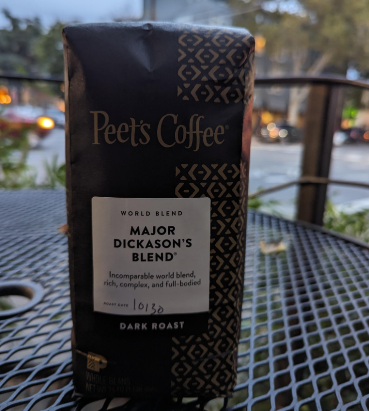 A bag of Peet’s Coffee Major Dickason’s Blend, World Blend, Dark Roast, sits on an outdoor table. The label describes it as rich, complex, and full-bodied. Blurred outdoor scenery is in the background.