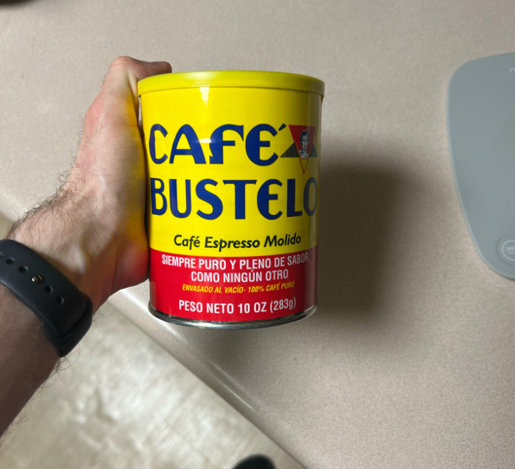 A hand holds a yellow and red can of Café Bustelo espresso coffee, labeled "Café Espresso Molido" with a net weight of 10 oz (283g), against a beige countertop background.