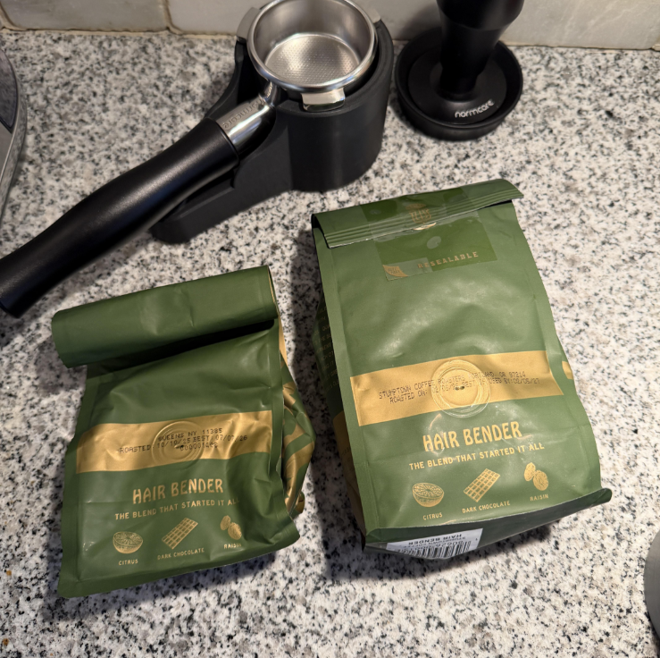 Two green bags of Hair Bender coffee sit on a speckled countertop next to a portafilter and a coffee tamper. The bags are labeled with the flavor notes: citrus, dark chocolate, and caramel.