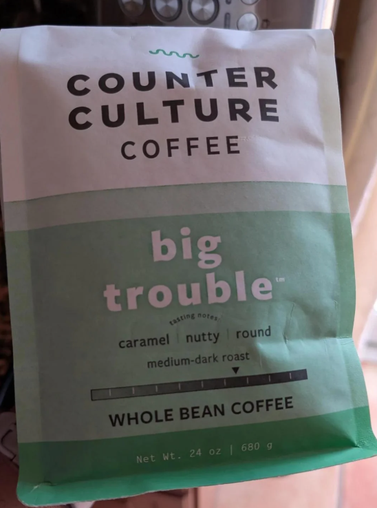 A bag of Counter Culture Coffee labeled "big trouble," described as caramel, nutty, and round, medium-dark roast, whole bean coffee. The bag is green and white and contains 24 oz (680 g).
