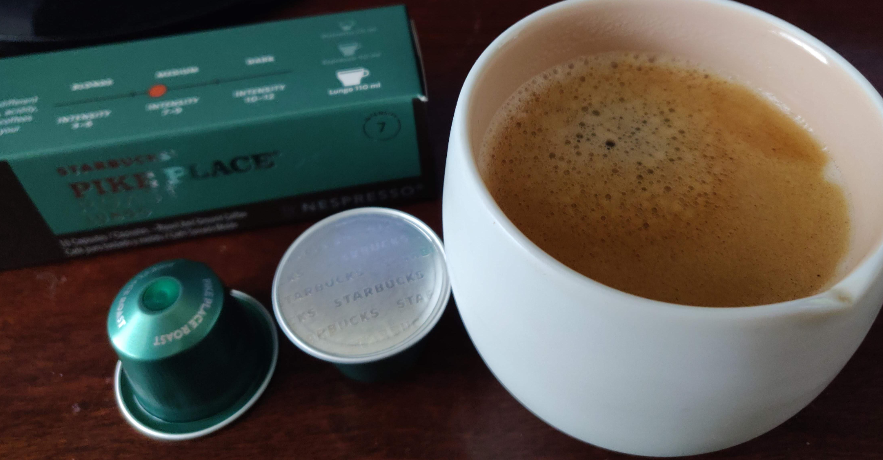 A cup of coffee with crema on top sits next to two green Nespresso coffee capsules and a Starbucks Pike Place Nespresso coffee box.