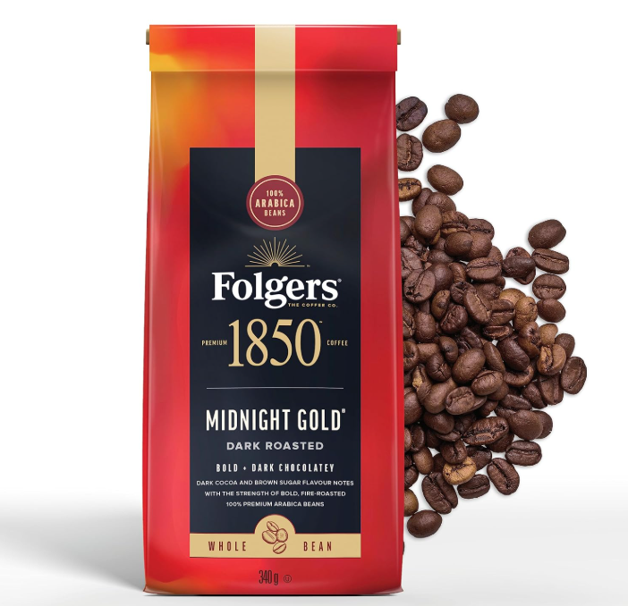 A bag of Folgers 1850 Coffee, Midnight Gold dark roasted whole bean, sits upright with coffee beans scattered to its right. The packaging features red, gold, and black colors with product details.