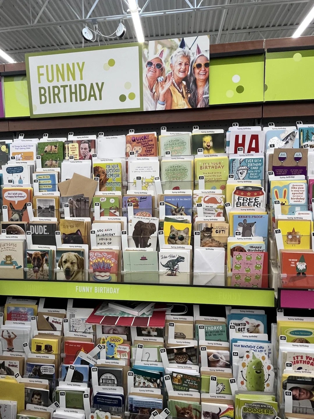 A store display of assorted funny birthday cards, featuring Walmart products, organized on several shelves under a "FUNNY BIRTHDAY" sign, with a photo of three smiling people in party hats above the display.