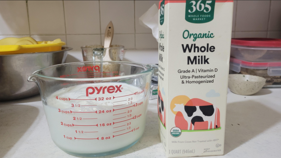 A carton of 365 Whole Foods Market organic whole milk stands next to a Pyrex measuring cup filled with milk on a kitchen counter with various utensils and dishes in the background.