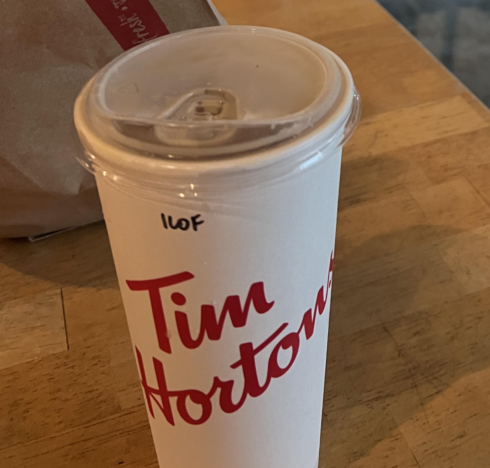 A Tim Hortons paper cup with a plastic lid sits on a wooden table. The cup has "110F" written on it in black marker. A brown paper bag is partially visible in the background.