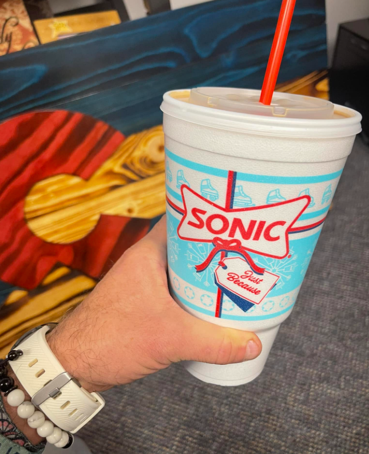 A hand holding a large Sonic Drive-In soft drink cup with a red straw, perfect for sipping fast food coffees. The cup features a blue and red design and "Sonic" branding. Abstract art with red, yellow, and blue colors decorates the background.