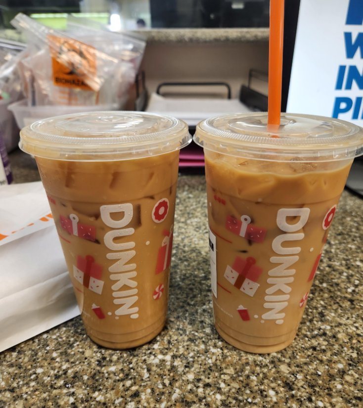 Two large fast food coffees from Dunkin', each with orange straws, sit on a speckled countertop. The cups feature colorful holiday-themed designs, while snacks and office supplies can be seen in the background.
