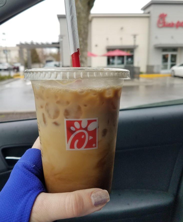 A hand holds an iced coffee with a straw in a clear cup featuring the Chick-fil-A logo, inside a car, with a Chick-fil-A restaurant visible through the window on a cloudy day—perfect for fans of fast food coffees.