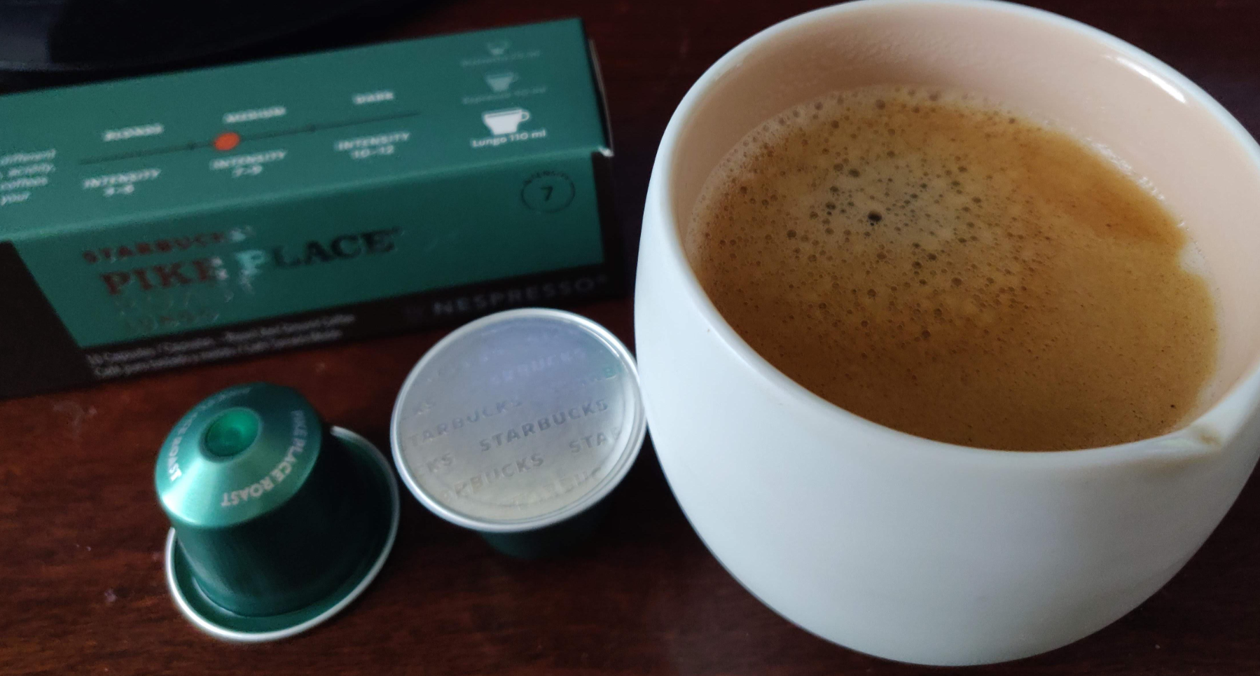A close-up of a cup of coffee with a creamy layer of crema, next to Starbucks Pike Place Nespresso capsules and their green packaging box on a wooden surface—perfect for those who prefer fast food coffees at home.