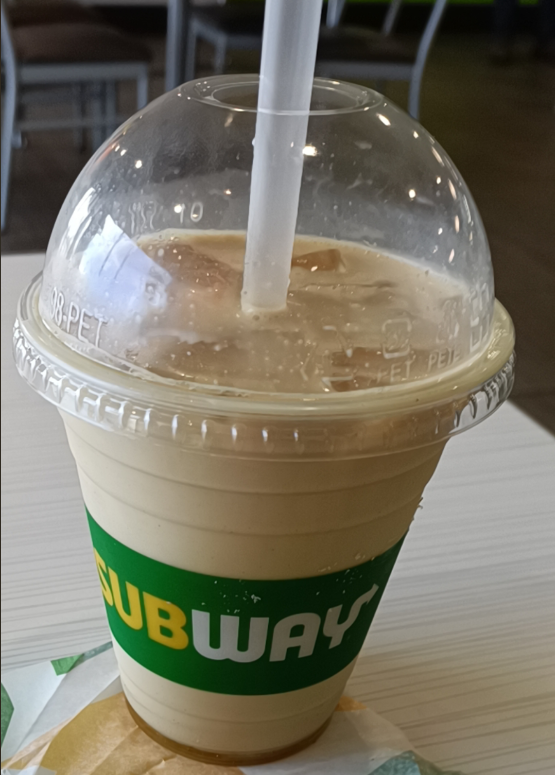 A plastic cup with a dome lid and straw, filled with a light brown iced drink, sits on a table. Part of a Subway sandwich wrapper is visible, suggesting classic fast food coffees to go. "SUBWAY" is printed on the cup.