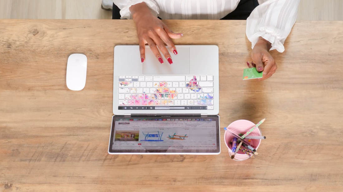 A person with red nail polish uses a laptop and holds a green credit card at a wooden desk, surrounded by a mouse, cup of pens, and an open website on a tablet—ready to shop online and hoping not to pay more for less.