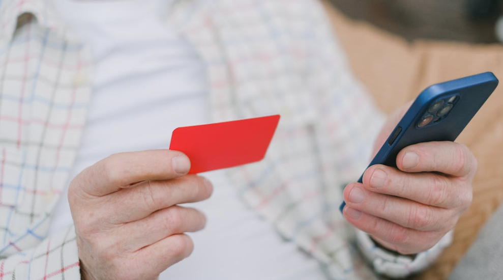 A person holding a red card in one hand and a blue smartphone in the other, wearing a plaid shirt and white t-shirt, seated on a beige surface—pondering whether to pay more for less.