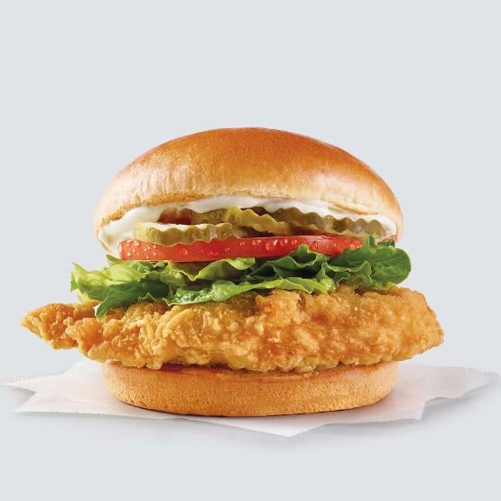 A crispy Chicken Sandwich with lettuce, tomato, pickles, and mayonnaise on a soft bun, displayed against a light background.