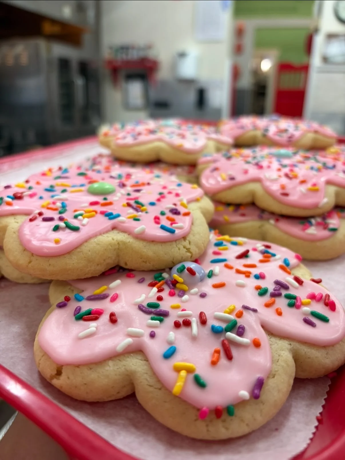 Heart-shaped sugar cookies with pink icing and colorful sprinkles are arranged on a red tray lined with parchment paper in a bakery or kitchen setting.