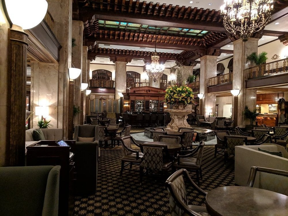 A luxurious hotel lobby reminiscent of Small-Town Luxury Hotels, with ornate chandeliers, marble columns, elegant furniture, a large central floral arrangement, and a high coffered ceiling with stained glass panels.