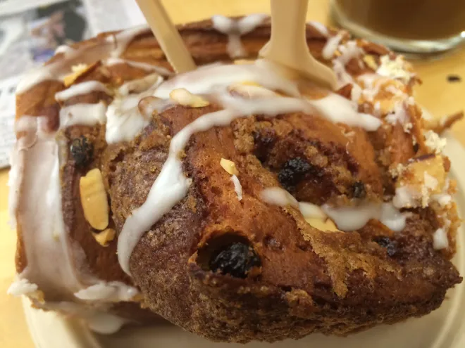 A close-up of a cinnamon roll topped with white icing and sliced almonds, with two wooden forks stuck into it. The cinnamon roll is on a plate, and part of a drink is visible in the background.