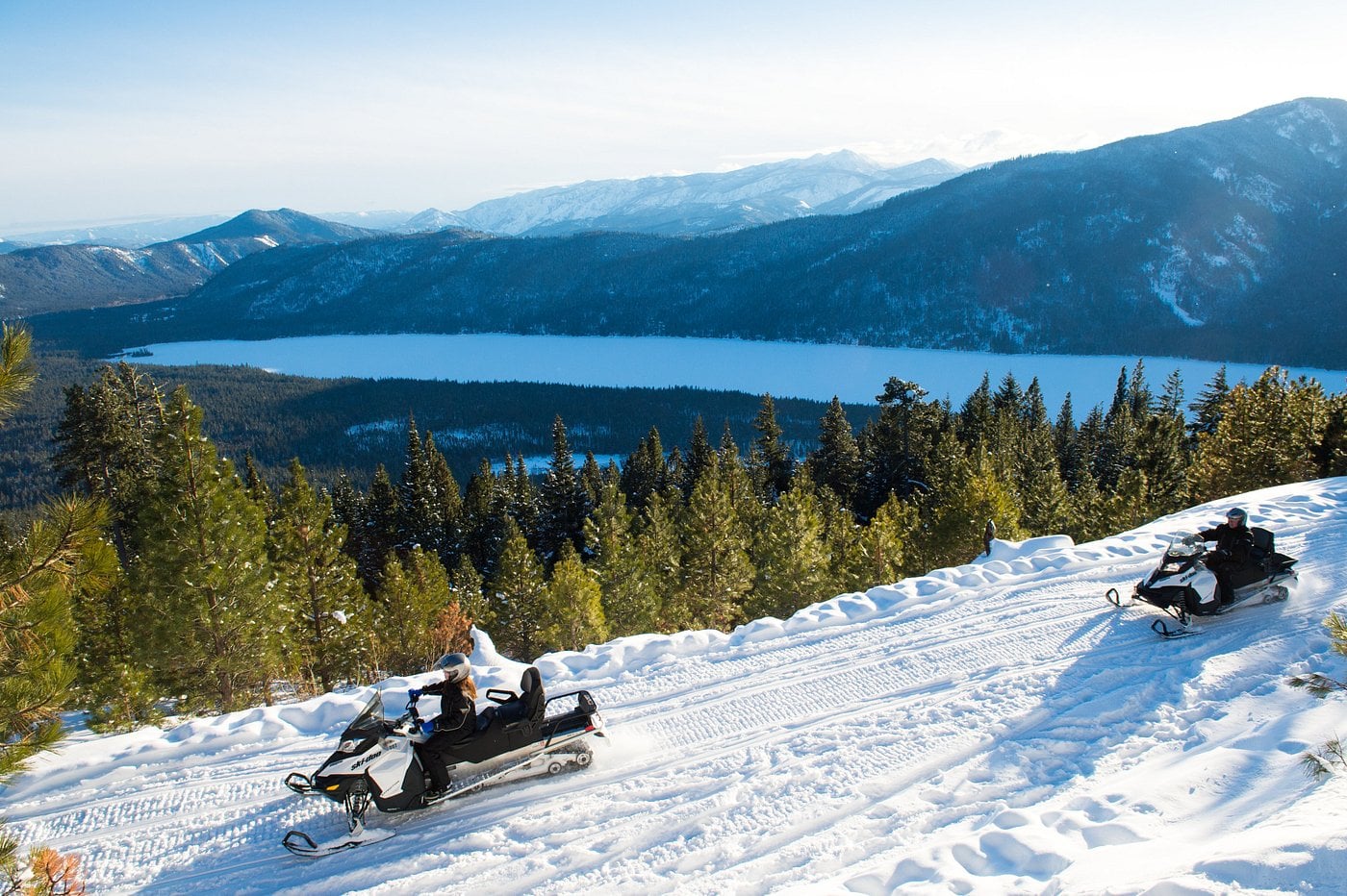 Two people ride snowmobiles on a snowy trail overlooking a lake and forested mountains under a clear sky, discovering the winter beauty found near unique American towns.