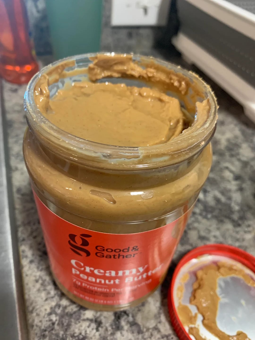 An open jar of Good & Gather creamy peanut butter, one of the best peanut butter brands, sits on a kitchen counter with some peanut butter on the rim and the red lid resting nearby.